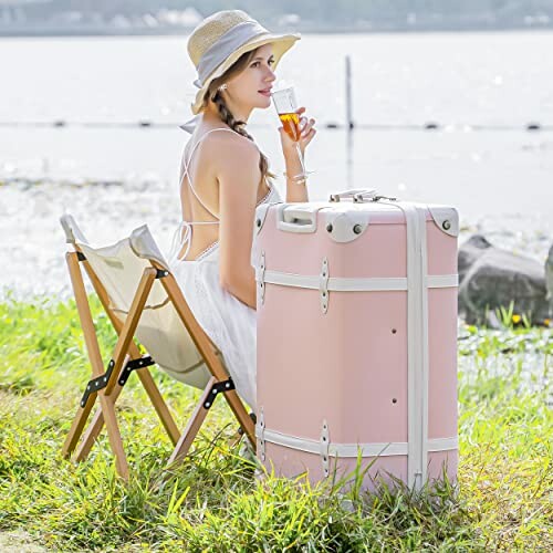 Woman in a hat sitting on a chair with a pink suitcase by a lake